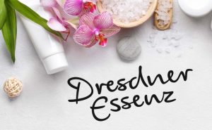Verweis auf Dresdner Essenz - im Fokus ist der Schriftzug "Dresdner Essenz" in der typischen Schriftart abgebildet. Der obere Teil des Bildes ist mit kleinen Steinen, Blättern, Meersalz und Orchideen dekoriert. Im oberen linken Bereich liegt eine weiße Handcreme Tube.