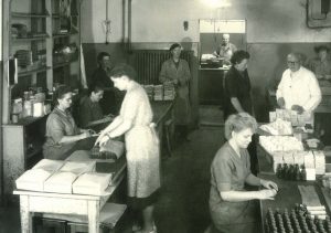 Ein historisches Bild der Li-iL GmbH. Auf dem Bild sind 10 Personen zu sehen, die verschiedene Tätigkeiten verrichten. Einige Personen befüllen und verpacken Produkte oder überwachen den gesamten Prozess. Das Bild ist in schwarz-weiß aufgenommen und zeigt den Ursprung der Arzneimittelherstellung bei der Li-iL GmbH.