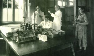 Vier Personen aus den 1910ern sind abgebildet, die Arzneimittel herstellen, eine Person hat bspw. ein Reagenzglas in der Hand. Das Bild ist in schwarz-weiß aufgenommen und zeigt den Ursprung der Arzneimittelherstellung bei der Li-iL GmbH.