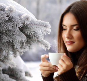 Eine Frau genießt mit geschlossenen Augen den Winter. Sie steht neben einem mit Schnee bedeckten Nadelbaum und hat ein dampfendes, heißes Getränk in der Hand.