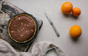 Ein frisch gebackener Kuchen steht in einer Backform auf einem Backblech. Auf dem Kuchen sind Mandelblätter und die Mandelblätter liegen auch auf dem Tisch verteilt. Neben dem Backblech liegt ein Küchentuch, ein Behälter mit Milch, ein Messer und einige Orangen.