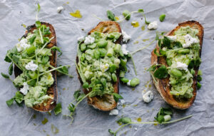 Auf einem Tisch liegen drei geröstete Brote nebeneinander. Die Brote sind belegt mit Guacamole, Kräutern, Lauchzwiebeln, Edamame und Frischkäse. Die Brote sind zusätzlich mit Olivenöl betröpfelt.