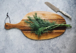 Ein Holzbrett liegt auf einem Tisch. Auf dem Holzbrett liegt ein Bund Dill und daneben liegt ein großes, scharfes Messer.